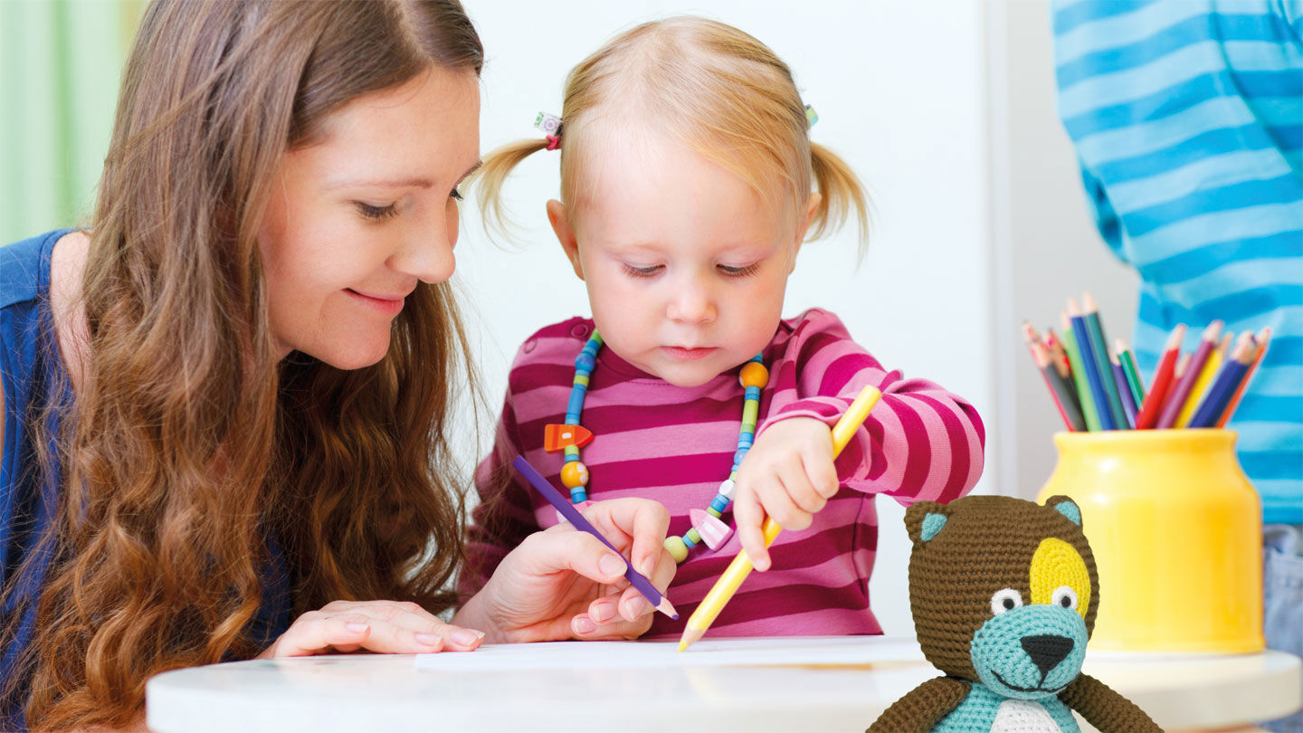 En mamma och ett litet barn ritar med kritor tillsammans. Nallen Tryggve sitter i förgrunden.