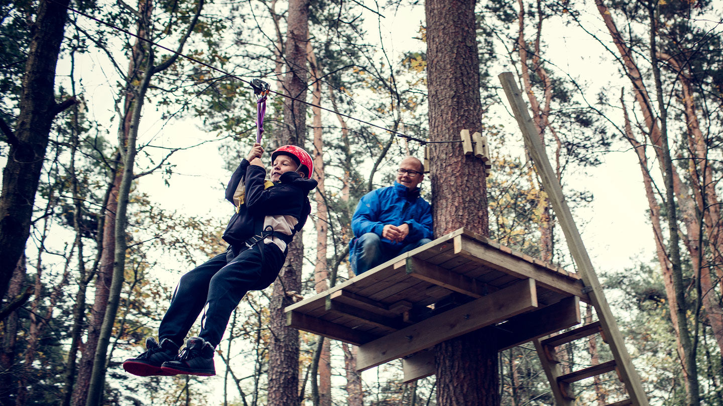 Ett barn åker linbana medan en vuxen tittar på.