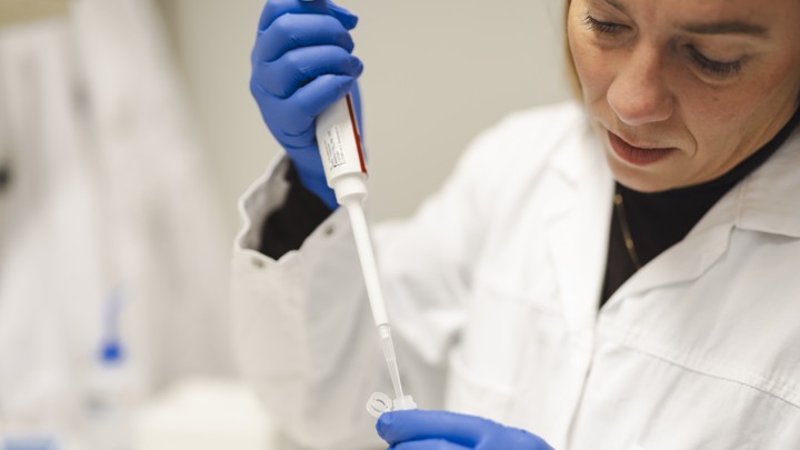 En kvinna i labbrock och bl&aring; handskar h&aring;ller i ett r&ouml;r